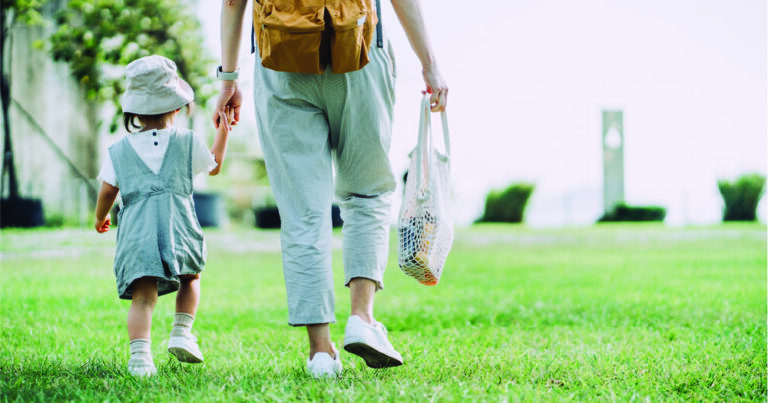 Parent holding hands with their child while walking on the grass and carrying a reusable bag