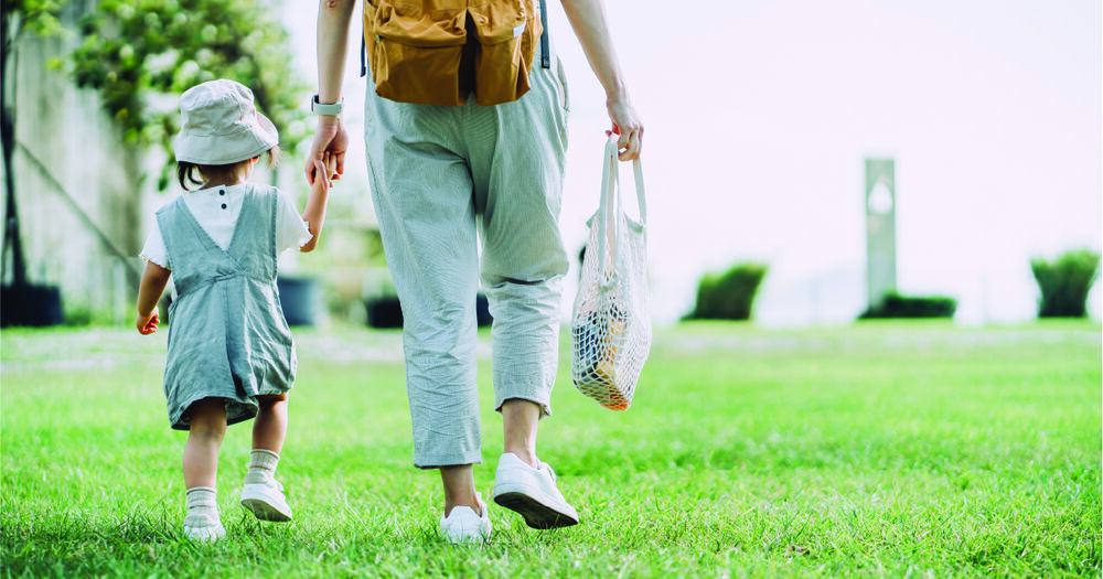 Parent holding hands with their child while walking on the grass and carrying a reusable bag