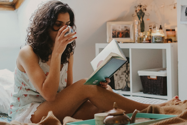 Woman drinking from a mug and reading a book on her bed